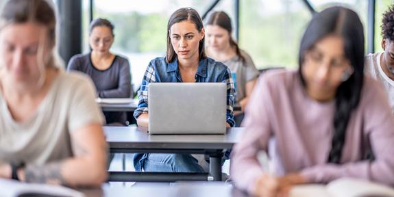 A female student sitting at a desk using a laptop in a classroom.
