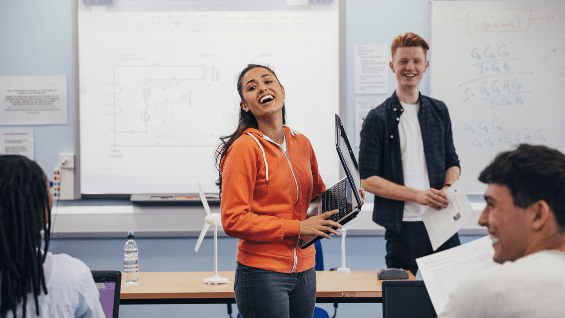 A young woman laughs and carries a laptop as she prepares to present her work to the class.