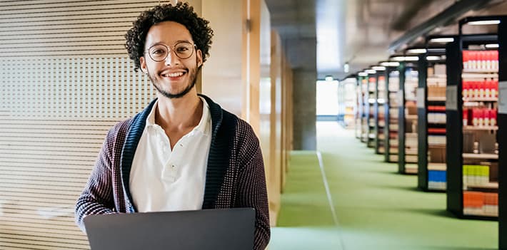 A librarian smiles at the camera while using his laptop in a library.