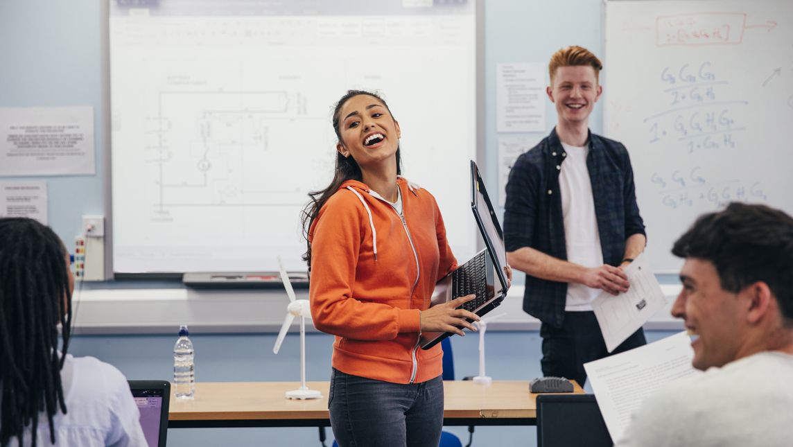 A young woman laughs and carries a laptop as she prepares to present her work to the class.