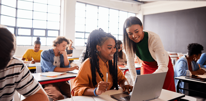 Smiling teacher talking with a student using a laptop during a classroom lesson